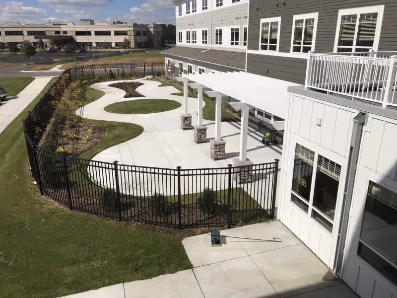 Outdoor patio area at Mill City Senior Living featuring a concrete walkway with curved design, a covered pergola supported by stone pillars, fenced garden beds with plants, and a multi-story building with windows and balconies in the background.