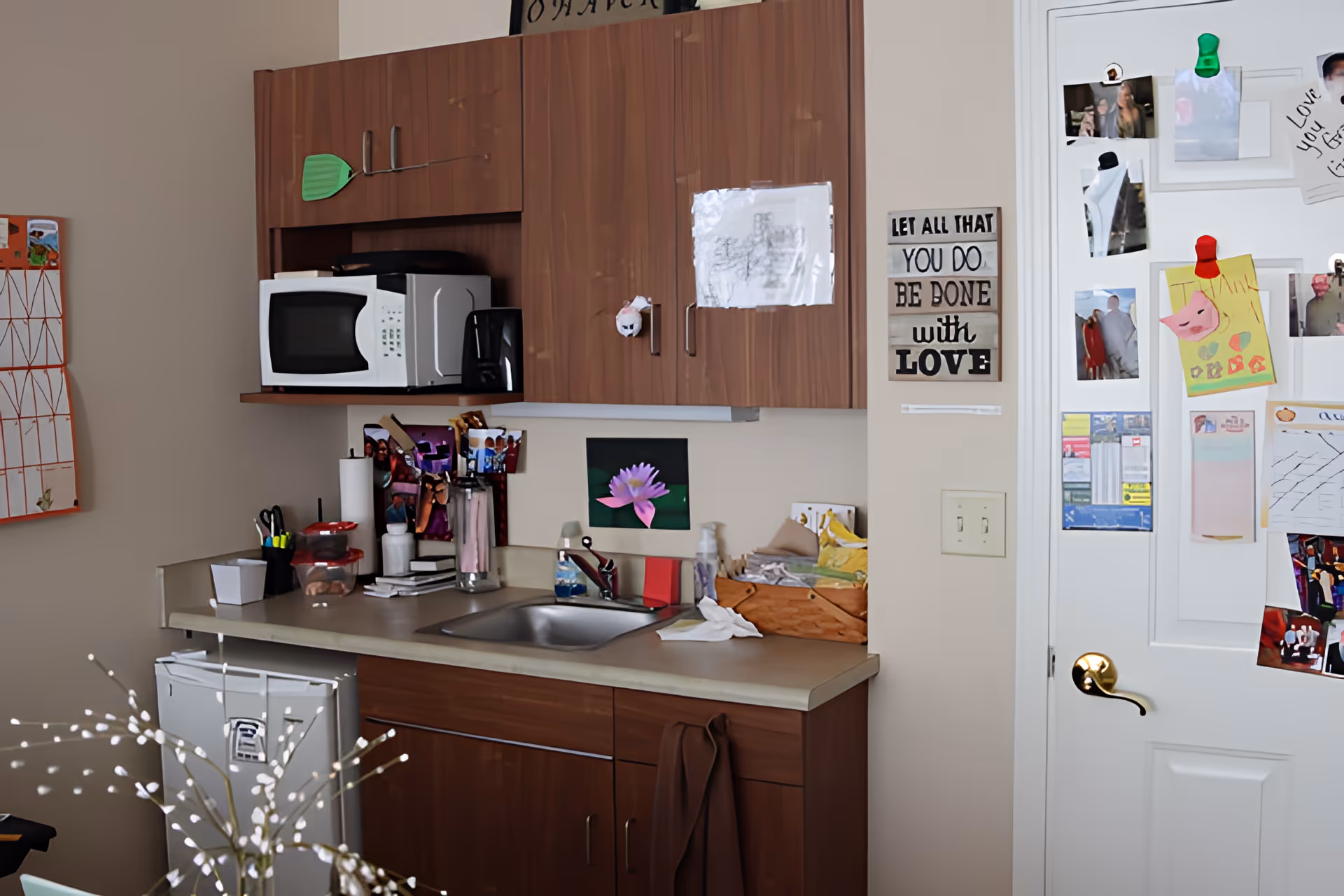 A small kitchen area with wooden cabinets, a microwave, a coffee maker, and a sink. The countertop has various items including a paper towel roll, containers, and a basket. The white door to the right is covered with photos, notes, and magnets. A sign on the wall reads 'LET ALL THAT YOU DO BE DONE with LOVE.'