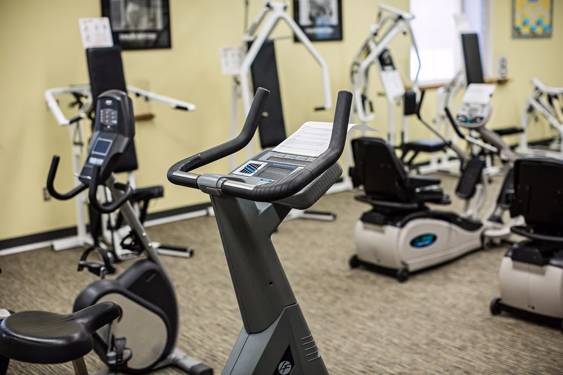 Indoor fitness room with various exercise equipment including stationary bikes and weight machines, set against yellow walls and carpeted floor.