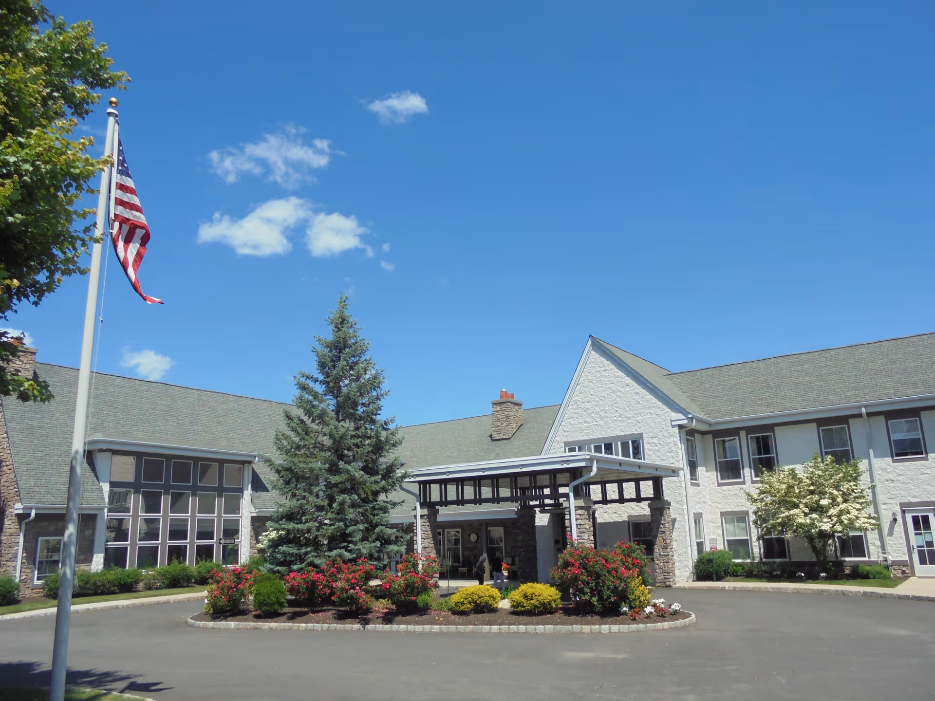 Exterior view of Independence Manor Hunterdon, showing a large building with a covered entrance, landscaped garden with bushes and a tall evergreen tree, an American flag on a flagpole, and a clear blue sky with a few clouds.