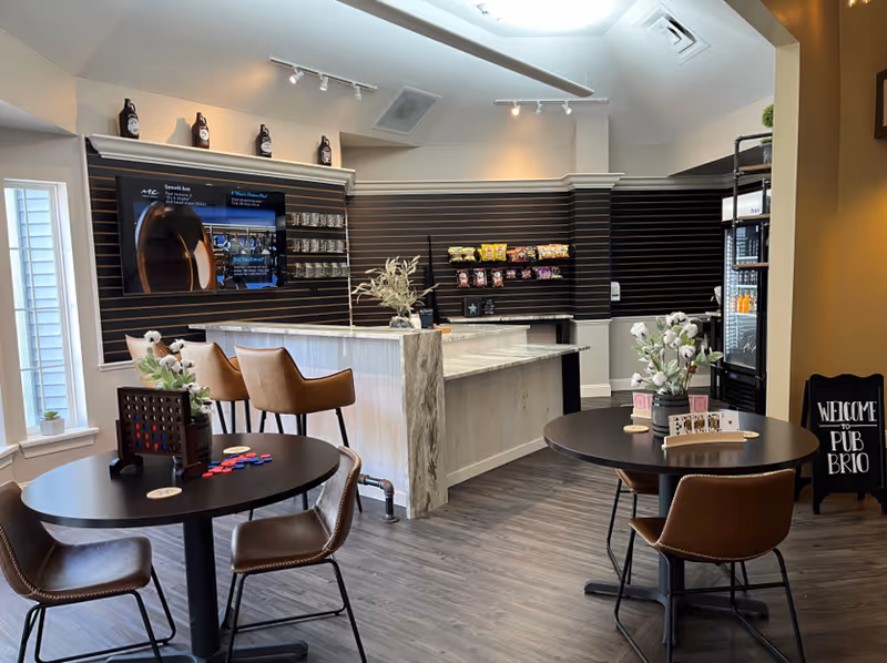 Interior view of a modern common area with two round tables and brown chairs, a counter with bar stools, shelves with snacks, a refrigerator, and a TV mounted on the wall. The space has wood flooring, a skylight, and decorative plants on the tables.