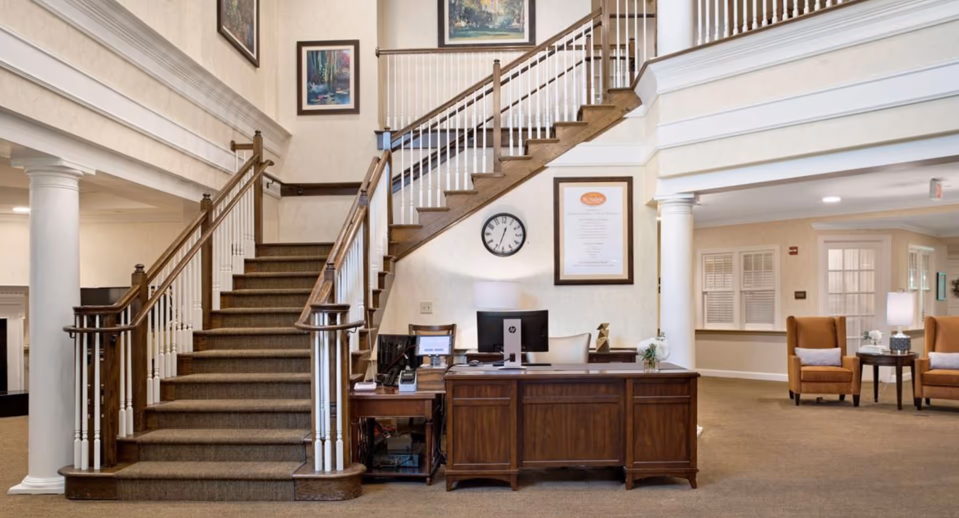 Interior view of a senior living facility lobby with a wooden staircase featuring white balusters, a wooden reception desk with a computer and lamp, a wall clock, framed artwork on the walls, and two brown armchairs with a small table and lamp in the background.