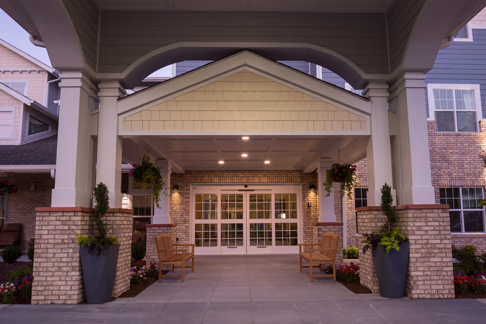 Entrance of a senior living facility with a covered drop-off area supported by brick and white columns. There are two wooden benches on either side of the entrance, large planters with greenery, hanging flower baskets, and double glass doors leading inside the building.