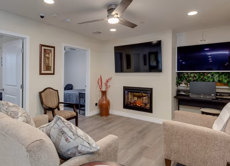 Cozy living room with sofas and armchairs facing a wall-mounted TV above a fireplace, with a desk area and doorway to a bedroom.
