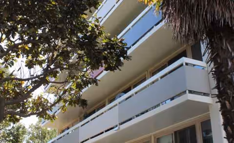 Multi-story senior living facility exterior with balconies, framed by trees in foreground.