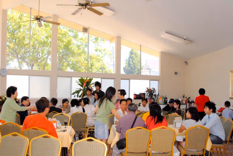 A bright dining room with many people seated at tables beneath tall windows and ceiling fans.