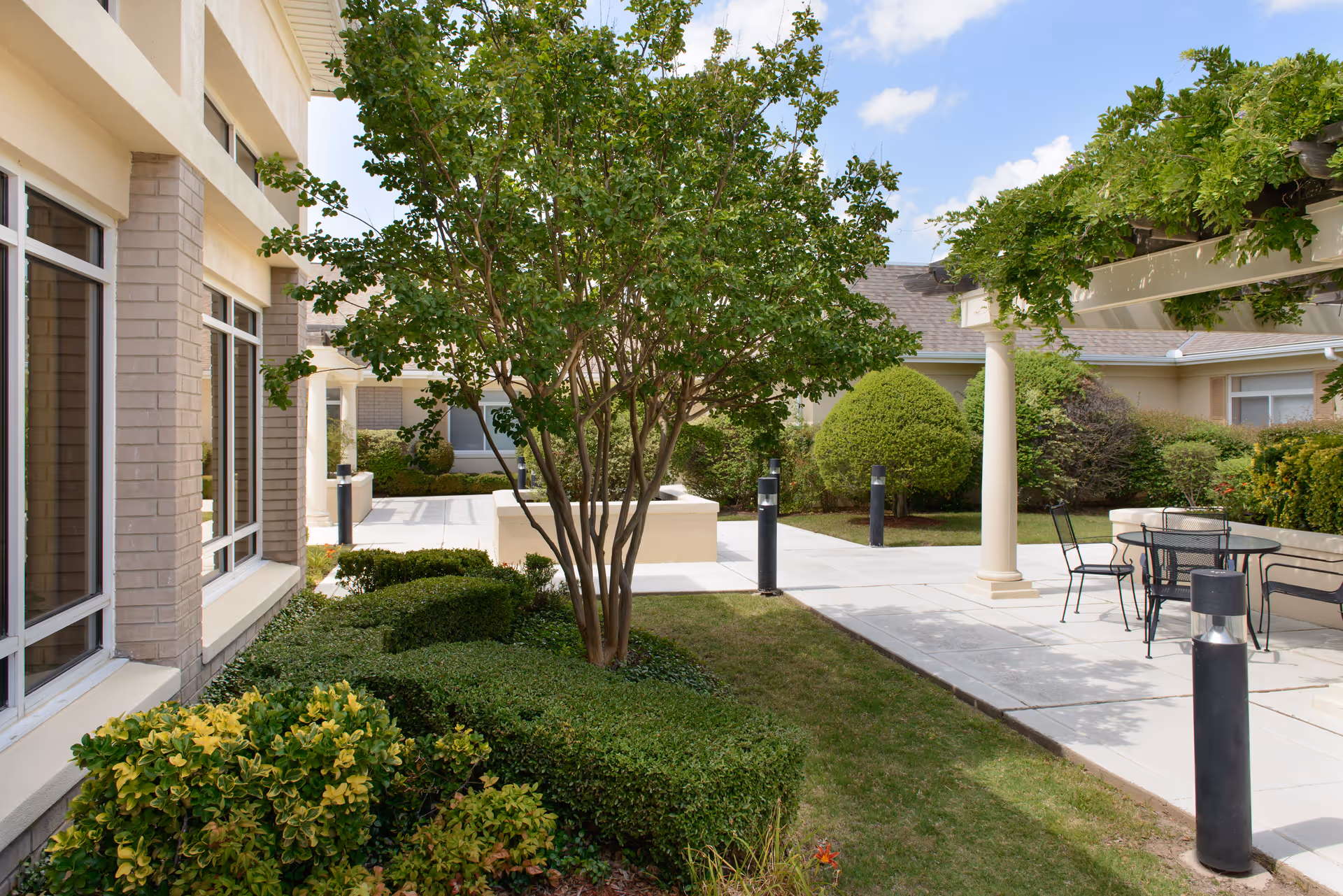 Outdoor courtyard area at Garden Terrace at Fort Worth featuring a paved walkway, green shrubs, a tree, and a seating area with a table and chairs under a pergola with greenery.