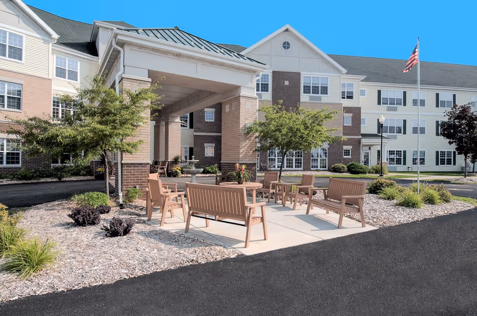 Outdoor seating area with wooden benches and chairs arranged around a table on a concrete patio in front of a multi-story senior living facility building. The building has a covered entrance, multiple windows, and is surrounded by landscaped plants and trees. An American flag is visible on a flagpole to the right.