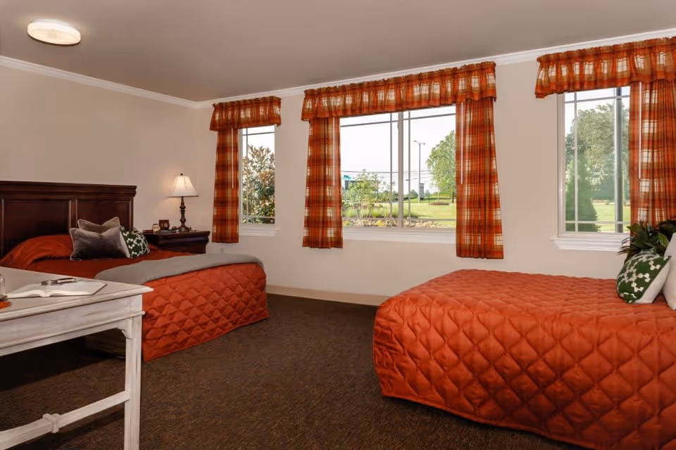 A bedroom with two beds covered in orange quilted bedspreads, each with decorative pillows. The room has three windows with orange plaid curtains, a wooden nightstand with a lamp, and a white table with an open book. Outside the windows, greenery and trees are visible.