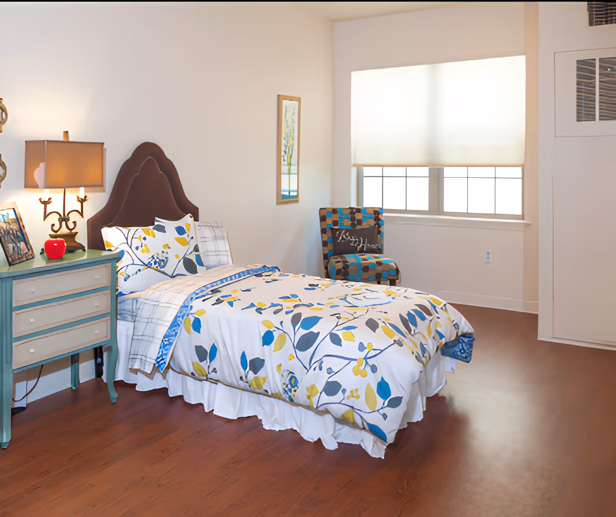 A bright bedroom with a single bed featuring a floral patterned comforter in blue, yellow, and gray colors. Next to the bed is a teal nightstand with a lamp, a red apple decoration, and framed pictures. A patterned chair with a pillow is placed near a window with a white blind. The room has wooden flooring and light-colored walls.