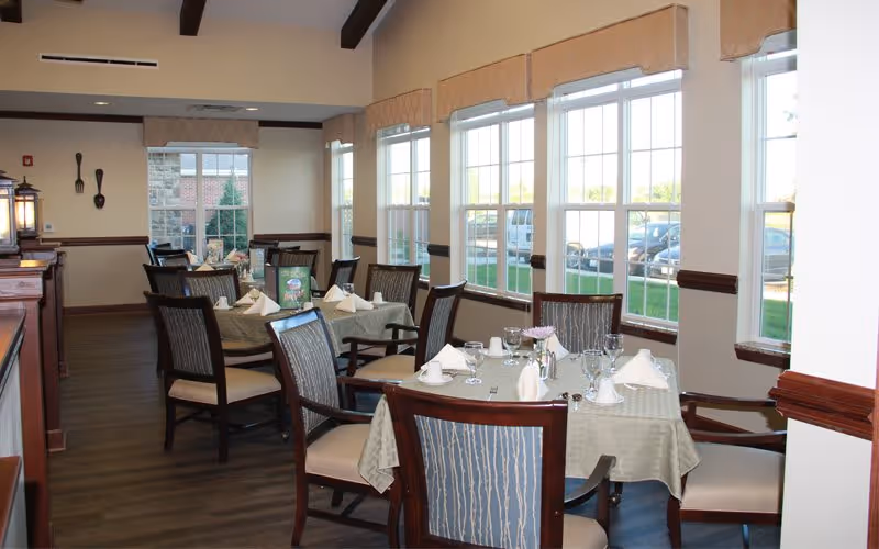 A bright dining room with several tables covered in light green tablecloths, each set with white napkins, glassware, and silverware. The room has large windows letting in natural light, wooden chairs with cushioned seats, and wooden beams on the ceiling.