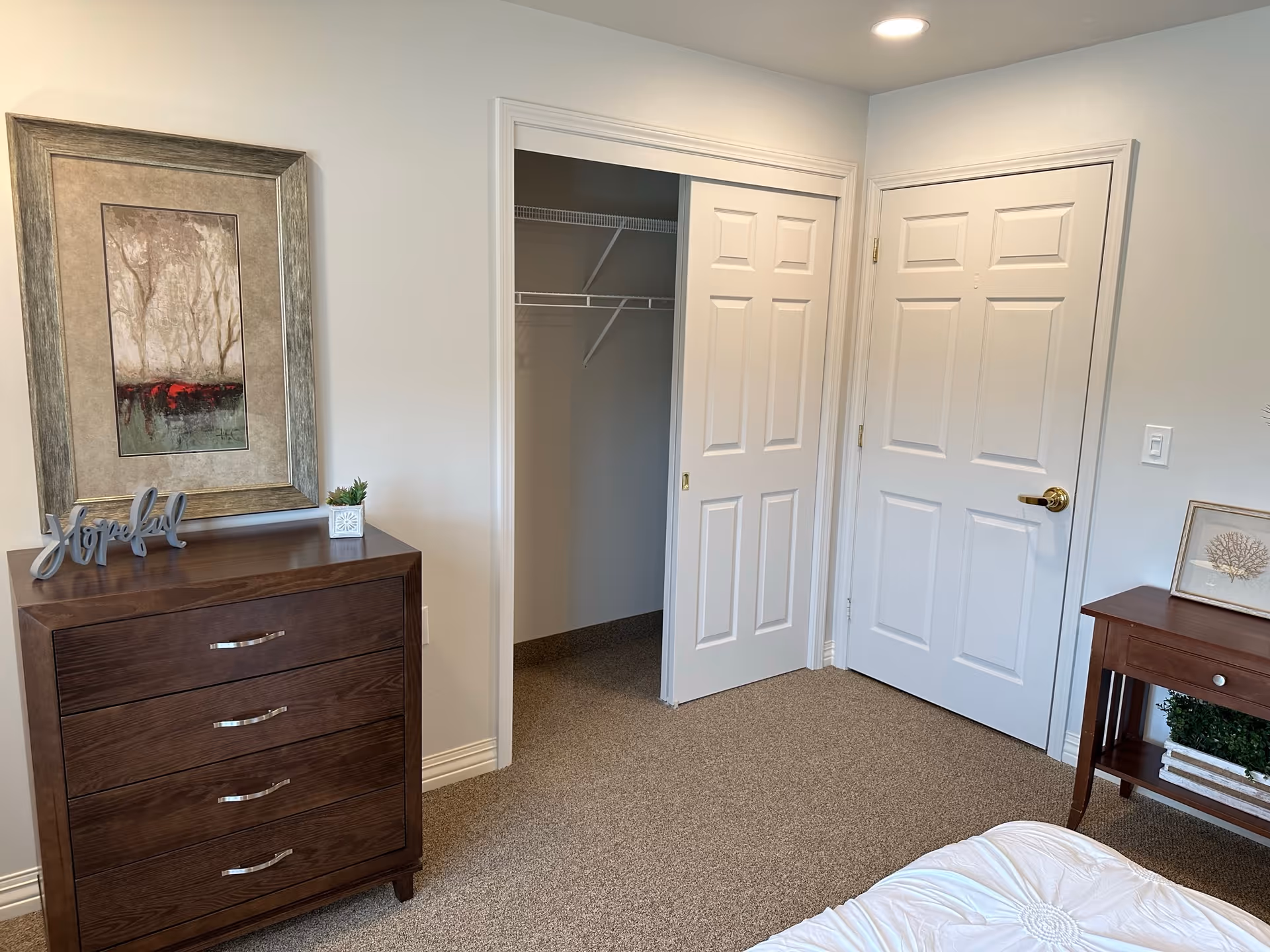 A bedroom corner featuring a wooden dresser with a decorative 'Joyful' sign and a small potted plant on top. Above the dresser is a framed artwork depicting trees. To the right, there is an open closet with white sliding doors and a white door with a gold handle. A small wooden side table with a framed picture and a green plant is next to the bed, which has a white comforter.