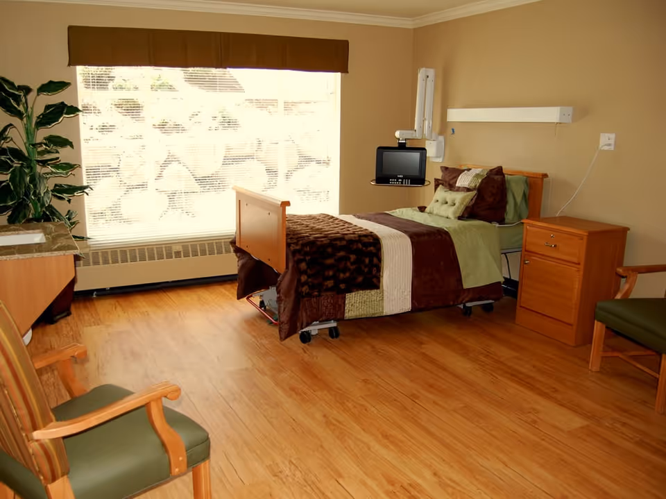 A bright, furnished patient bedroom with a single adjustable bed, bedside cabinet, chairs, and a large window.