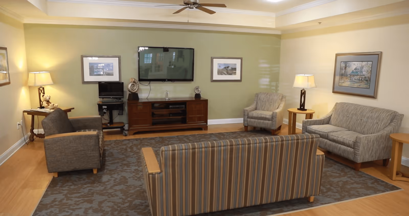 A cozy living room with a striped sofa in the foreground, two armchairs, and a loveseat arranged around a large area rug. The walls are painted in light green and beige tones, decorated with framed artwork. A flat-screen TV is mounted on the green wall above a wooden media console. Two table lamps provide warm lighting, and a ceiling fan is visible above.