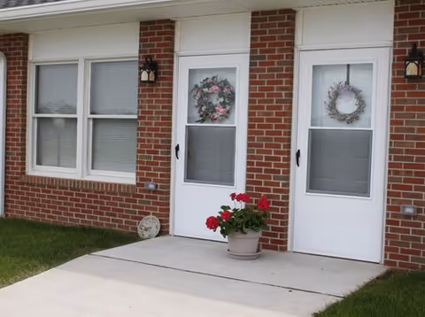 Exterior view of a brick building with two white doors, each decorated with a floral wreath. There is a potted plant with red flowers placed on the concrete walkway in front of the doors, and two windows with closed blinds to the left of the doors.