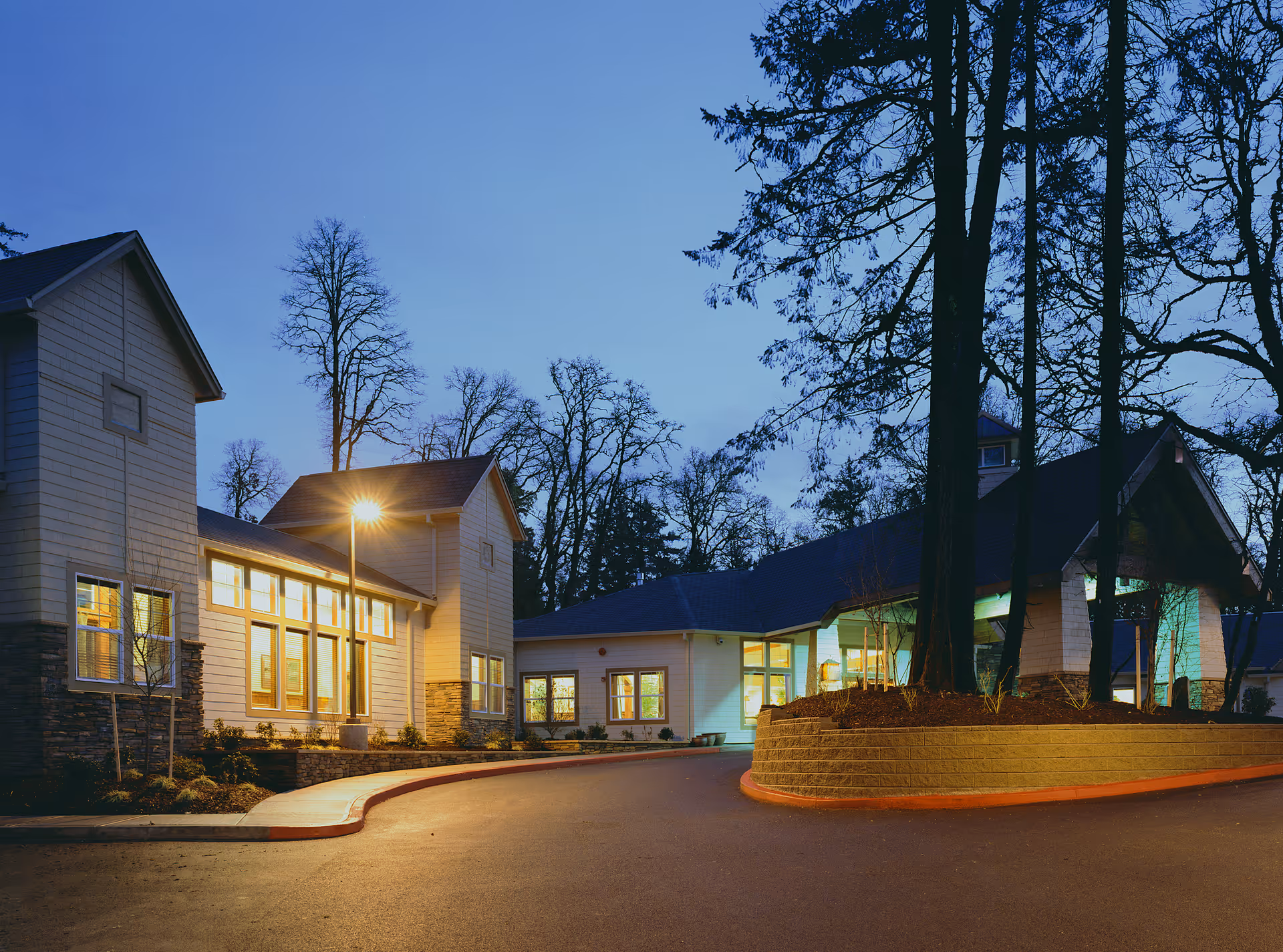 Dusk exterior view of a senior living facility entrance with illuminated windows, a curved driveway, and tall trees.