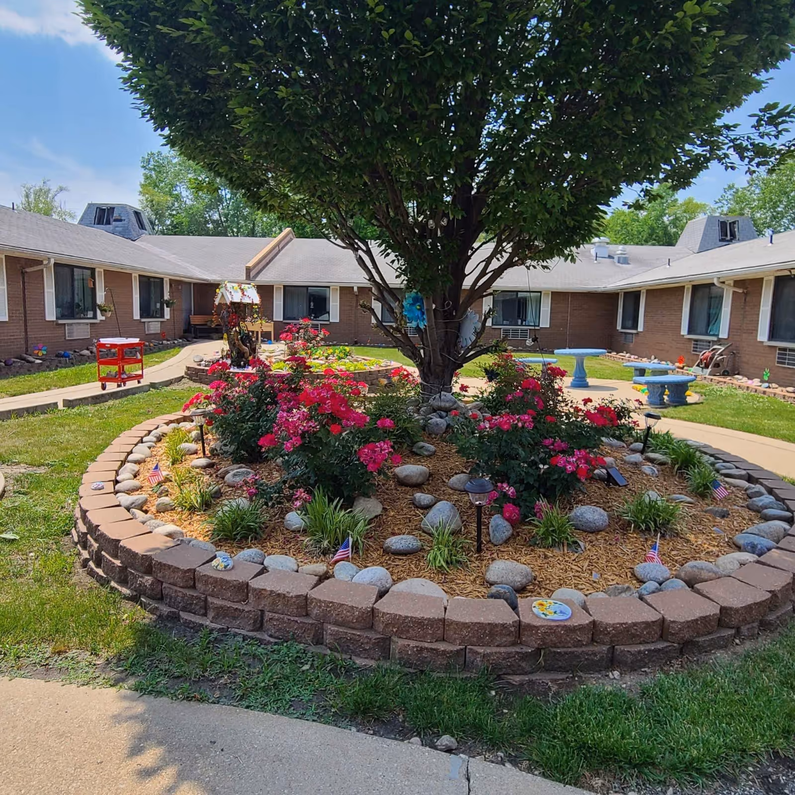 A landscaped courtyard garden at Carriage Square Rehab & Healthcare Center featuring a large tree surrounded by blooming pink flowers, rocks, and mulch bordered by a low brick wall. The courtyard is enclosed by a single-story brick building with windows and air conditioning units. There are paved walkways, a birdhouse, and blue stone benches in the background under a partly cloudy sky.