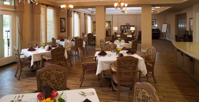 Bright, neatly arranged dining room with multiple white-clothed tables, patterned chairs, chandeliers, and a serving counter.
