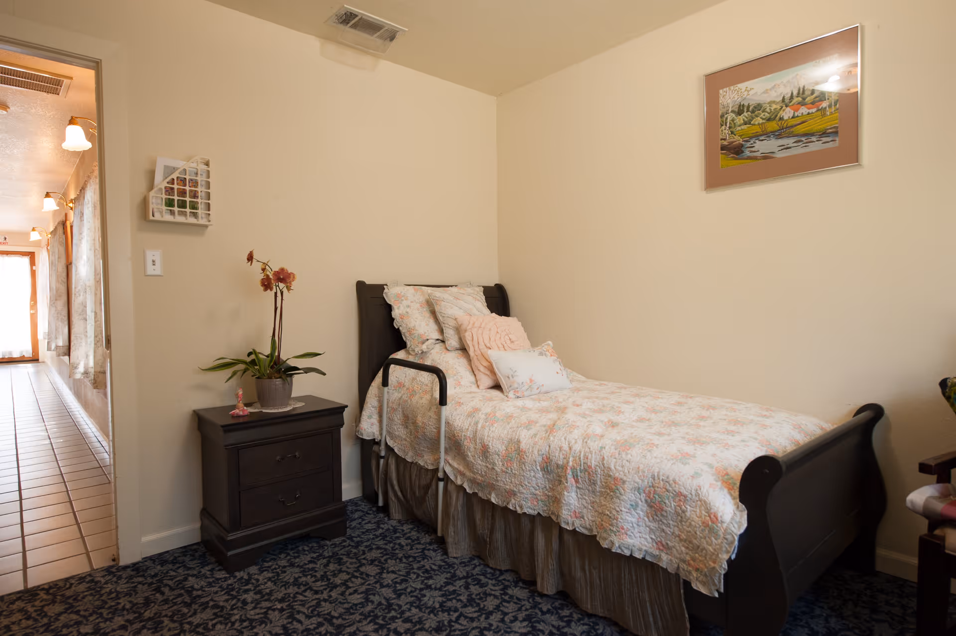 A small bedroom with a single bed covered in a floral quilt and several pillows. Next to the bed is a dark wooden nightstand with a potted orchid plant on top. The room has beige walls, a patterned carpet, and a framed landscape painting hanging above the bed. A hallway with tile flooring and wall lights is visible through an open door.