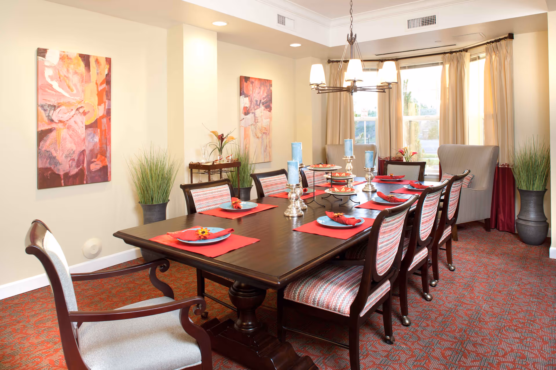 A well-lit dining room with a long dark wooden table set for eight people. Each place setting has a blue plate on a red placemat with a folded red napkin and a small flower. The chairs have patterned upholstery. The room features two abstract paintings on the wall, two tall potted plants, a chandelier with multiple lights, and large windows with beige curtains letting in natural light.