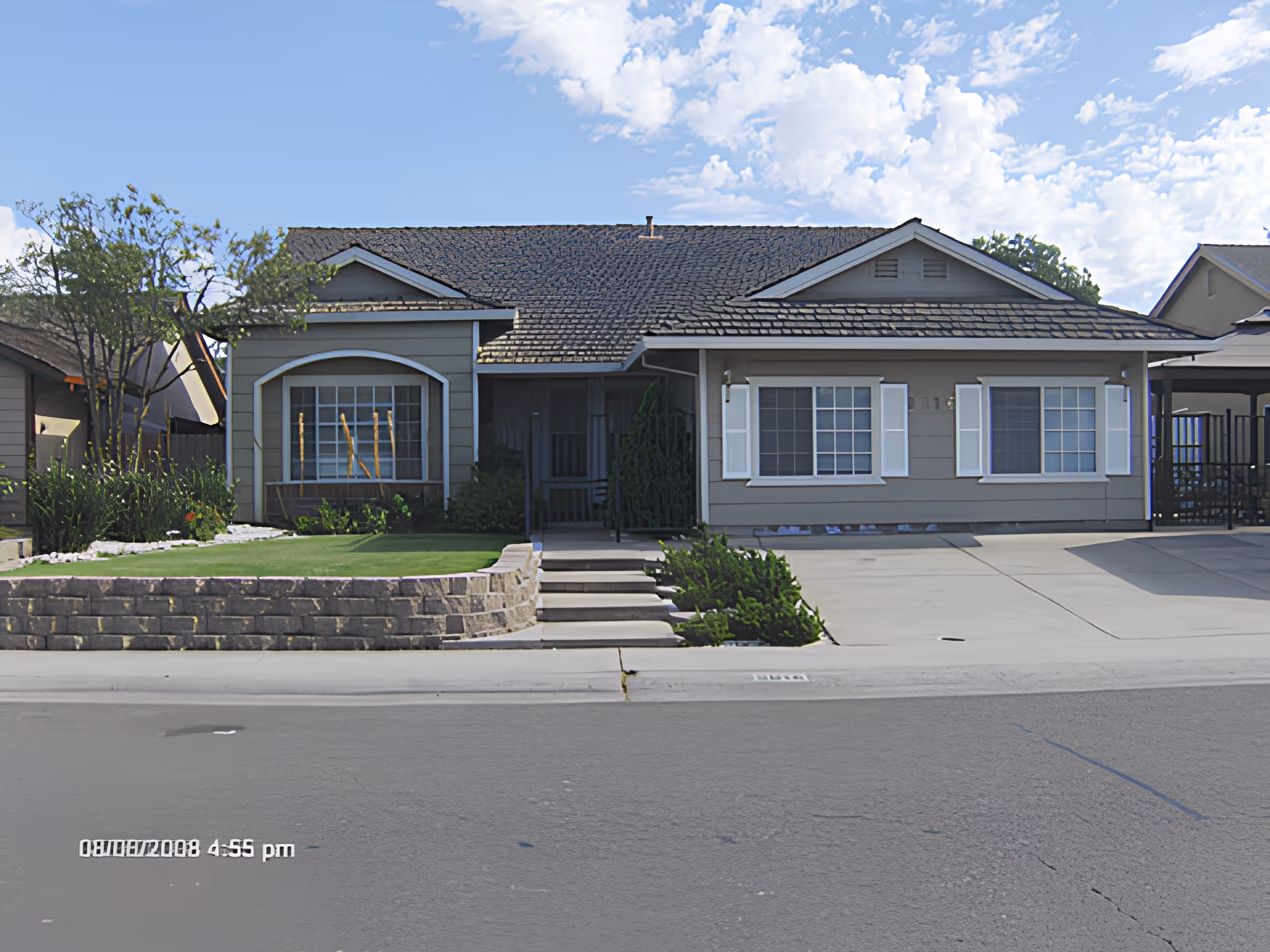 Single-story ranch-style house with a front porch, manicured lawn, low retaining wall and a driveway under a partly cloudy sky.