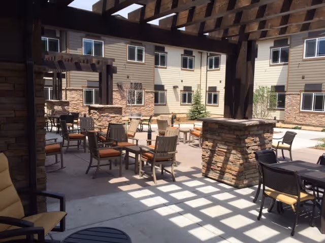 Outdoor patio area with multiple tables and chairs under a wooden pergola casting shadows on the concrete floor. The background shows a multi-story building with beige siding and stone accents, along with some small trees and shrubs.