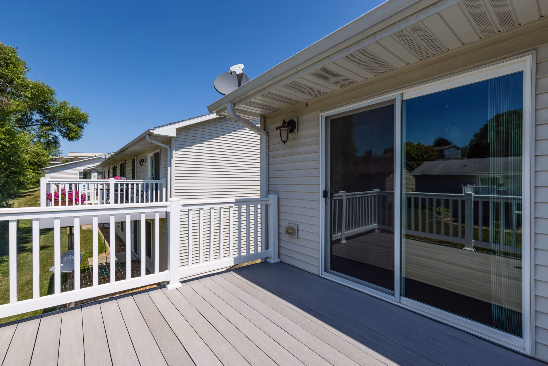 A second-floor balcony with white railing and sliding glass door on a light-colored vinyl-sided building overlooking a grassy yard and neighboring units.