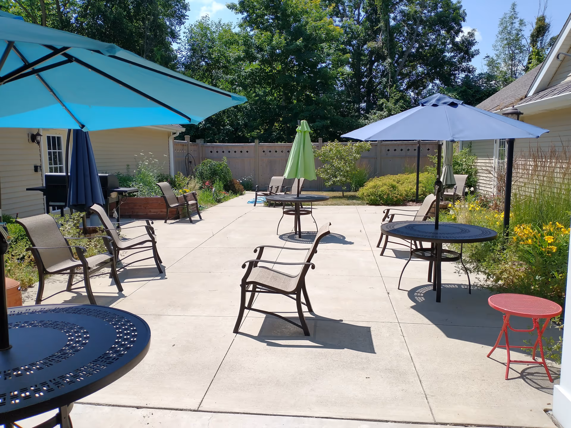 Outdoor patio area with several metal tables and chairs, each table shaded by large umbrellas in blue, green, and navy colors. The patio is surrounded by greenery, including bushes and trees, with a wooden fence in the background and beige buildings on either side.