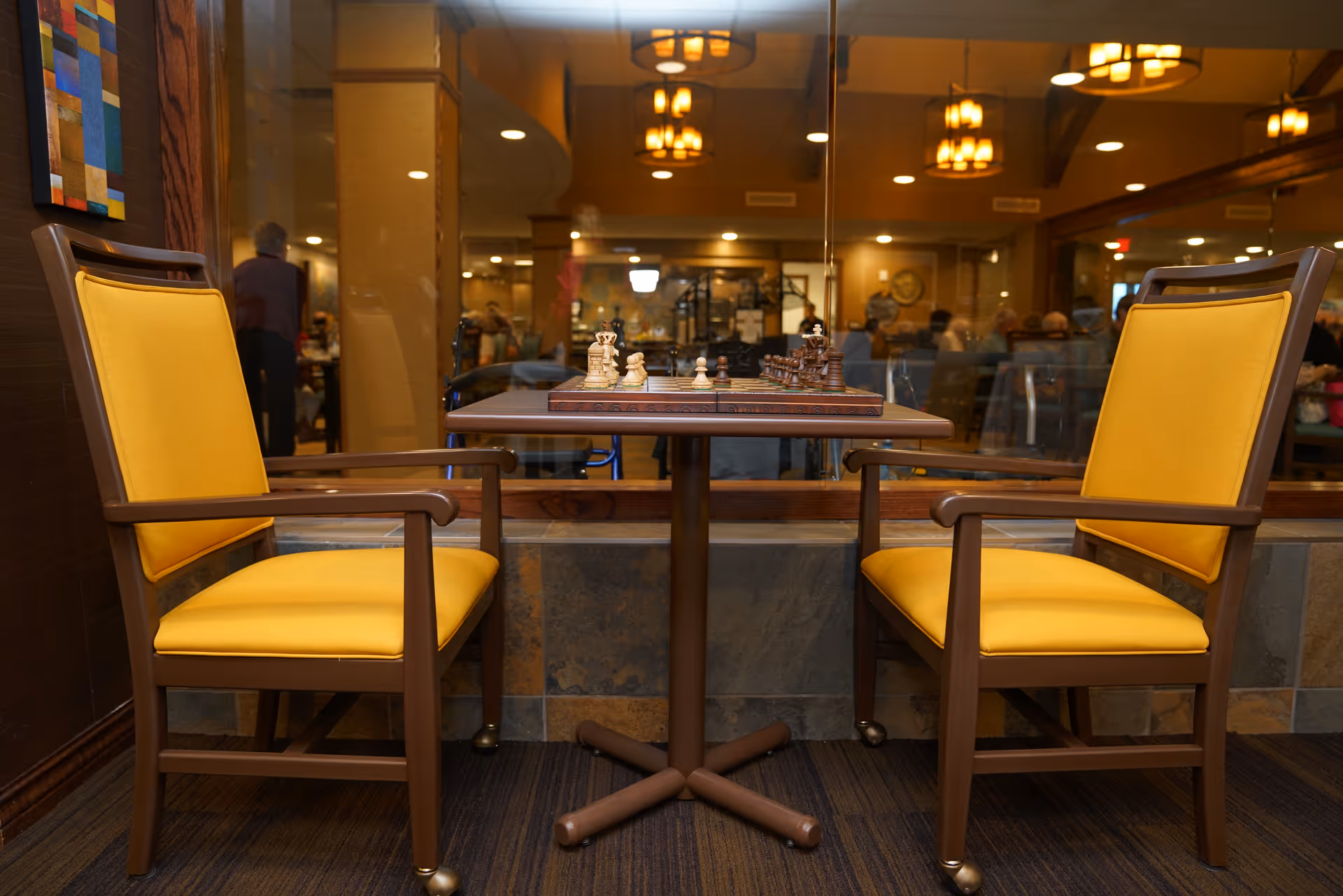 Two yellow-upholstered chairs face a small table with a chessboard in a retirement community common area.