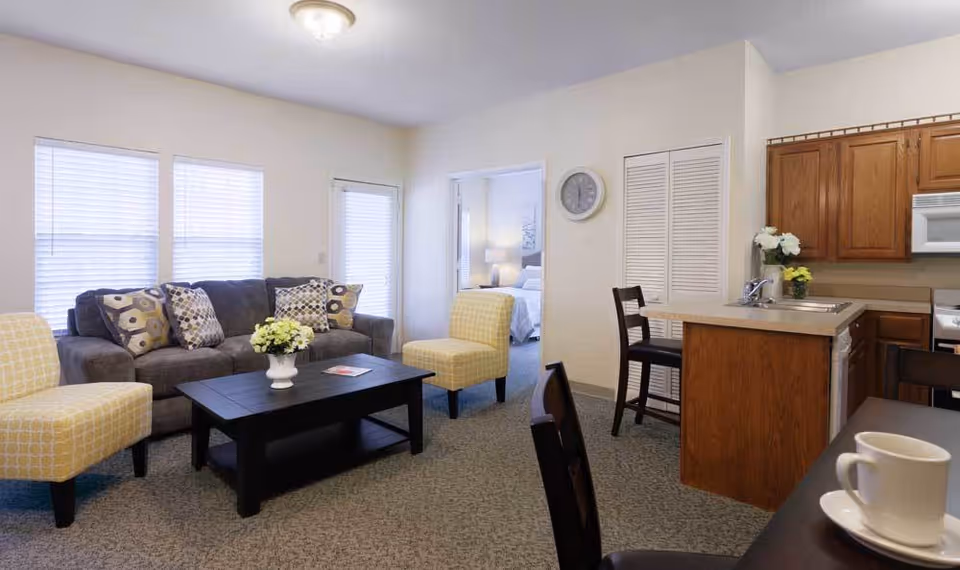 A cozy living room area with a gray sofa adorned with patterned pillows, two yellow patterned armchairs, and a black coffee table with a white vase of flowers. The room has large windows with blinds, a door with a window, and an open view into a bedroom with a bed and lamp. To the right is a kitchen area with wooden cabinets, a sink, and a microwave. A clock is mounted on the wall above a closed closet door.