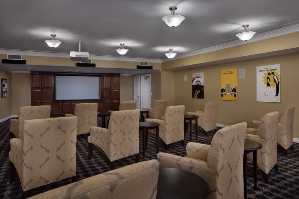 A cozy media room with multiple beige patterned armchairs arranged in rows facing a large flat screen TV mounted on a wooden cabinet. The room has beige walls adorned with colorful vintage-style posters, a patterned carpet, and ceiling lights evenly spaced across the ceiling.