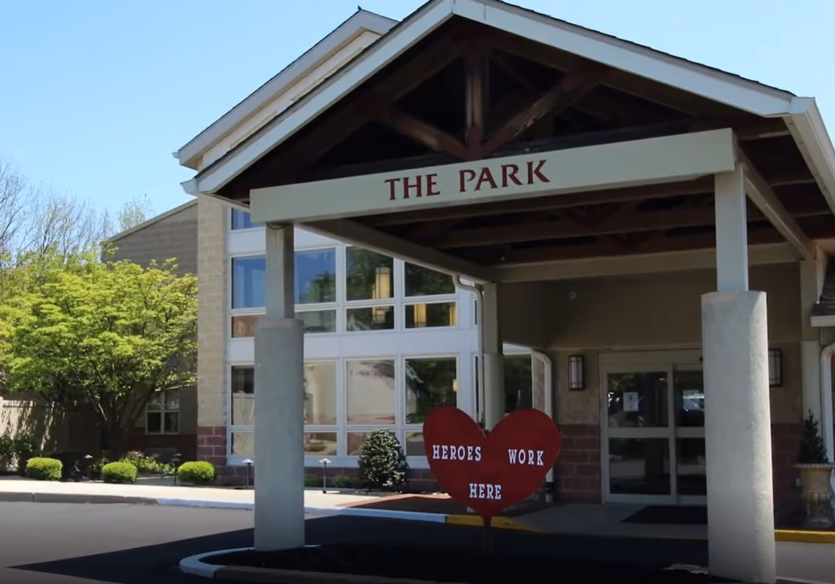 Entrance of a senior living facility named 'The Park' with a covered drop-off area supported by columns. In front of the entrance, there is a red heart-shaped sign that reads 'HEROES WORK HERE'. The building has large windows and is surrounded by greenery and trees.