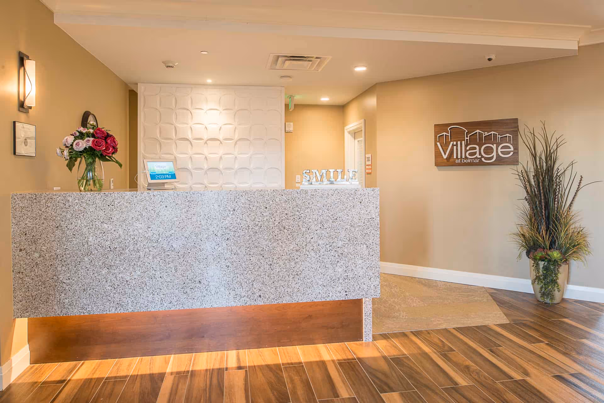 Reception area of Village at Belmar featuring a granite front desk with a vase of flowers on the left side, a decorative white wall panel behind the desk, a sign spelling SMILE on the desk, and a wooden Village at Belmar sign on the beige wall to the right next to a tall plant in a pot.
