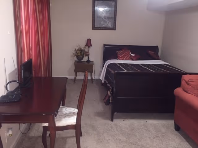 A bedroom with a dark wooden bed frame and a bed made with a dark patterned blanket and red pillows. To the left of the bed is a small wooden nightstand with a red lamp and a decorative plant. A wooden desk with a chair is positioned against the left wall, with a small TV on the desk. Red curtains cover the window behind the desk. A red upholstered chair is partially visible on the right side of the image. The room has beige walls and carpeted floor.