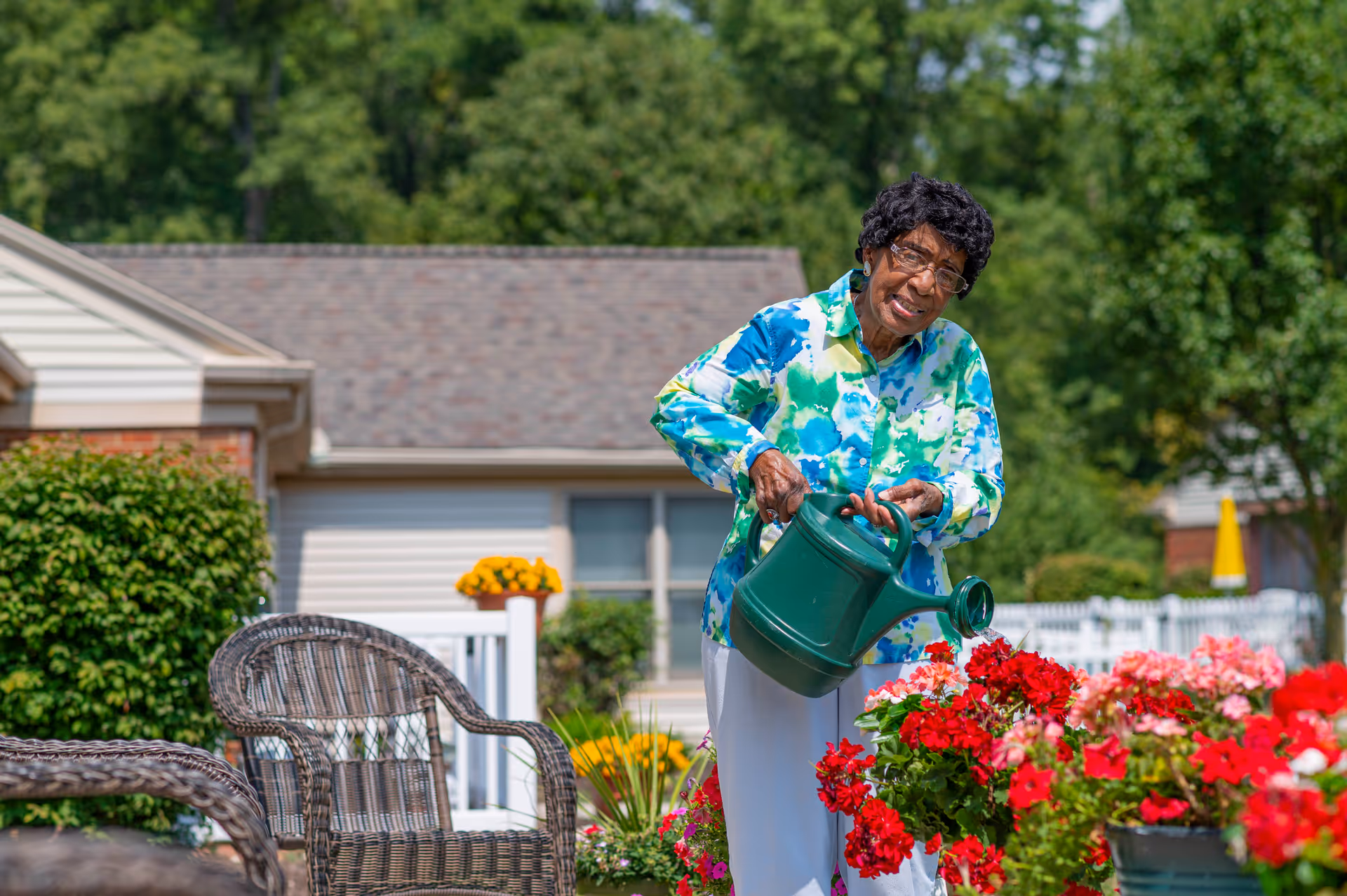 An elderly woman wearing glasses and a colorful blue and green blouse is watering red and pink flowers in a garden outside a residential building. There are wicker chairs and greenery around her, with a white fence and trees in the background.