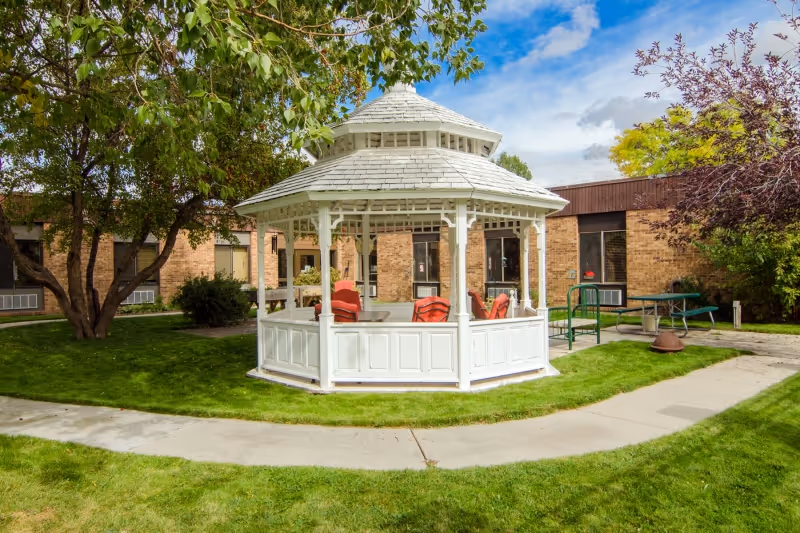 A white wooden gazebo with a shingled roof situated on a grassy lawn surrounded by a curved concrete pathway. The gazebo contains several red cushioned chairs. In the background, there are brick buildings and trees with green and purple foliage under a partly cloudy blue sky.