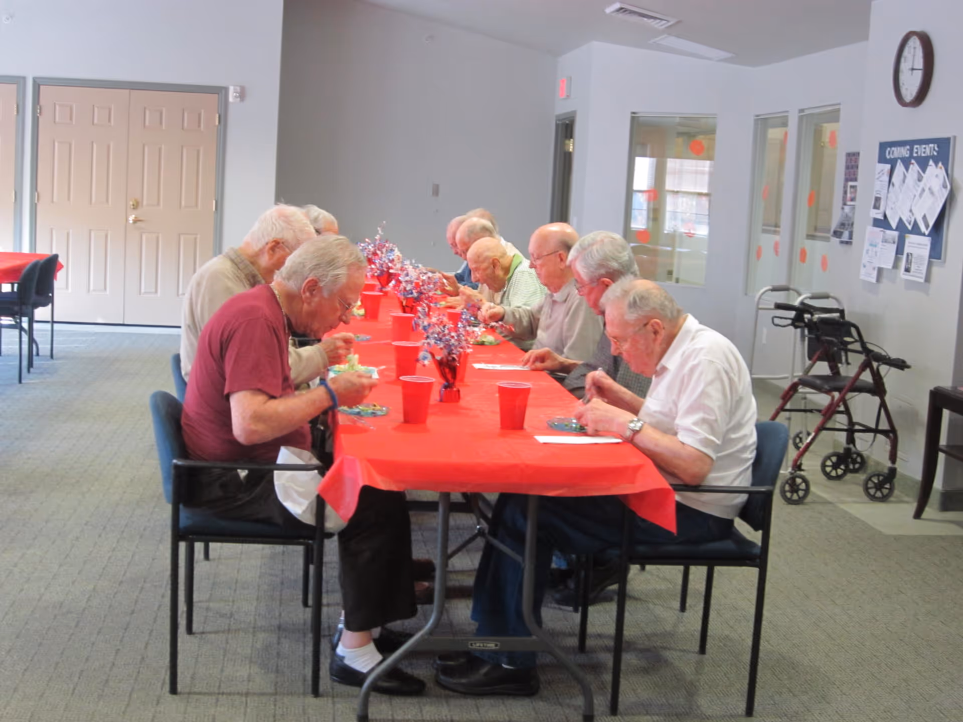 A group of elderly men sitting at a long table covered with a red tablecloth, eating a meal together in a communal dining area. The room has light-colored walls, a clock on the wall, a bulletin board with notices, and a walker nearby.