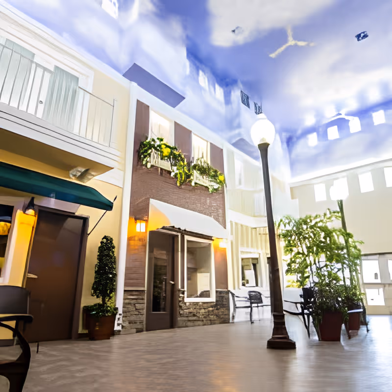 Indoor courtyard area of a senior living facility designed to resemble an outdoor street scene with building facades, windows with flower boxes, a street lamp, potted plants, benches, and a high ceiling painted to look like a blue sky with clouds.
