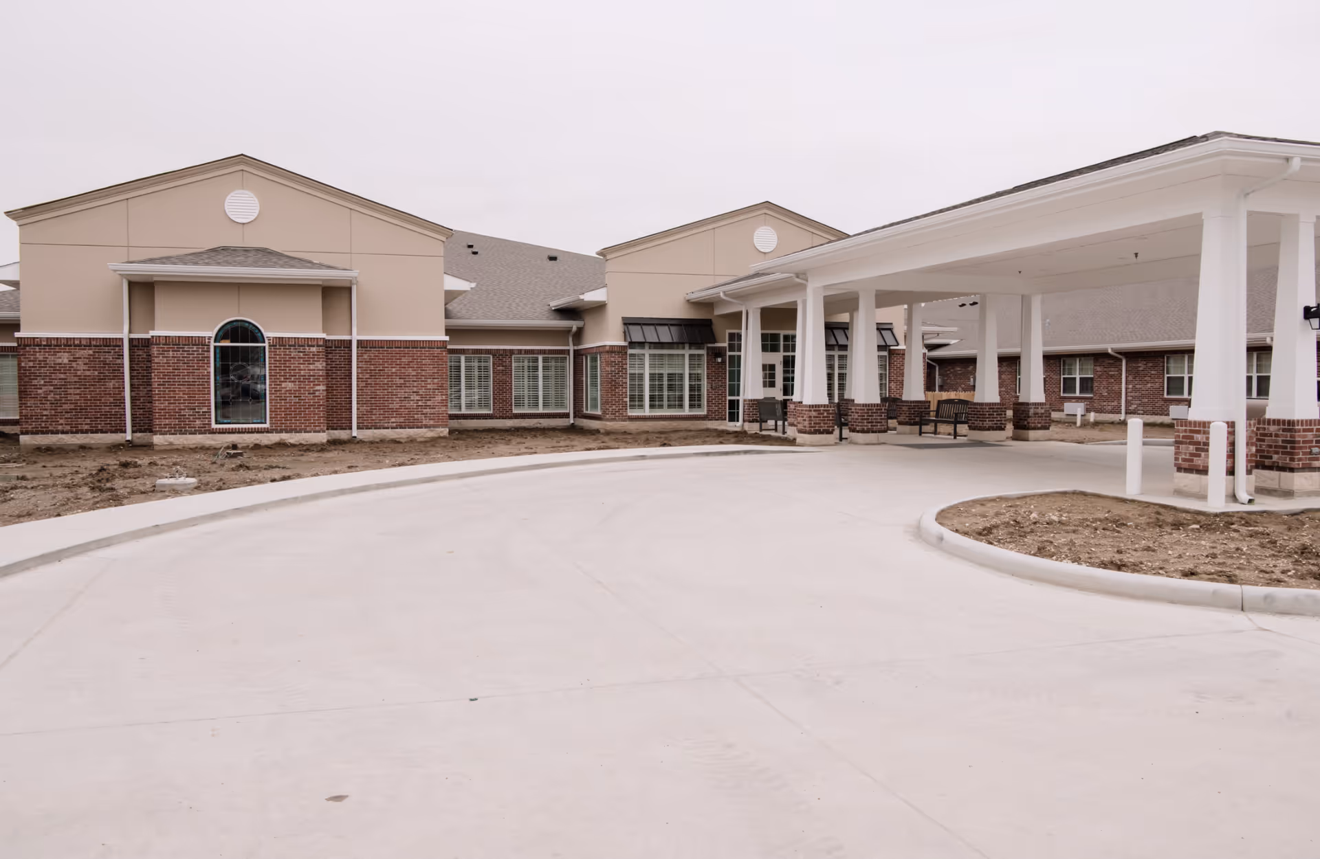 Exterior view of a senior living facility building with beige and brick walls, large windows, and a covered entrance with white pillars. The driveway is wide and curves around the entrance area.
