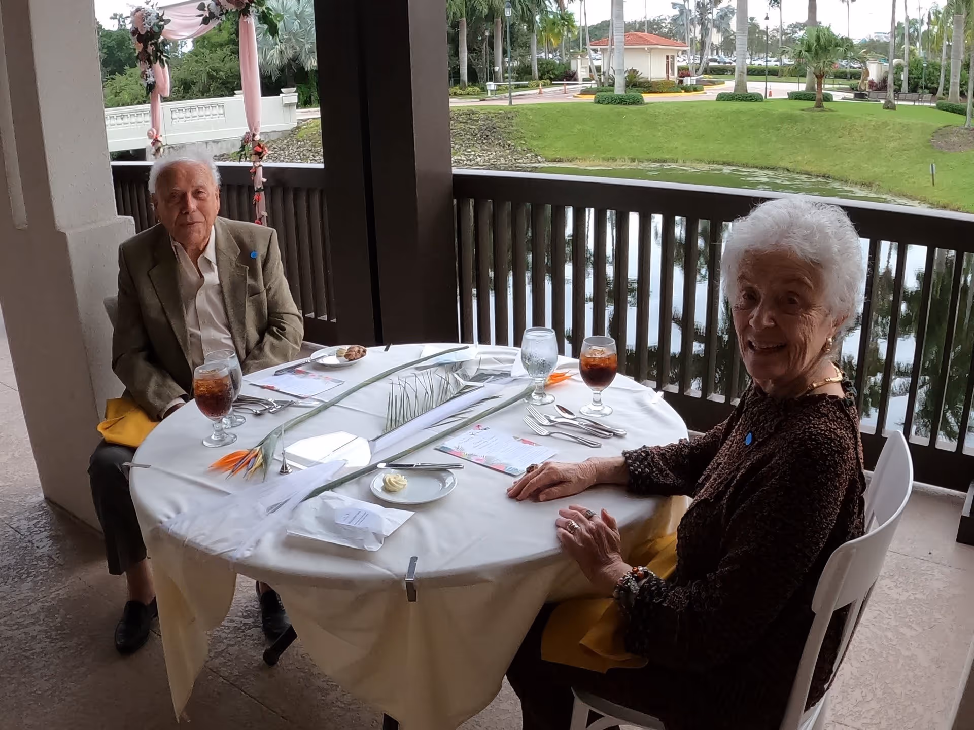 Two elderly individuals sitting at a round table covered with a white tablecloth on a covered patio overlooking a pond and green lawn. The table is set with glasses of iced tea, menus, utensils, and a small plate with butter. The background shows palm trees and a building with a red roof.