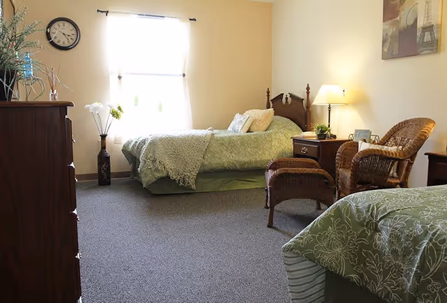 A cozy bedroom with two beds covered in green bedding. There is a wicker chair with a matching ottoman next to a nightstand with a lamp and a small plant. A dresser with decorative items and a wall clock is on the left side. A window with sheer curtains lets in natural light.