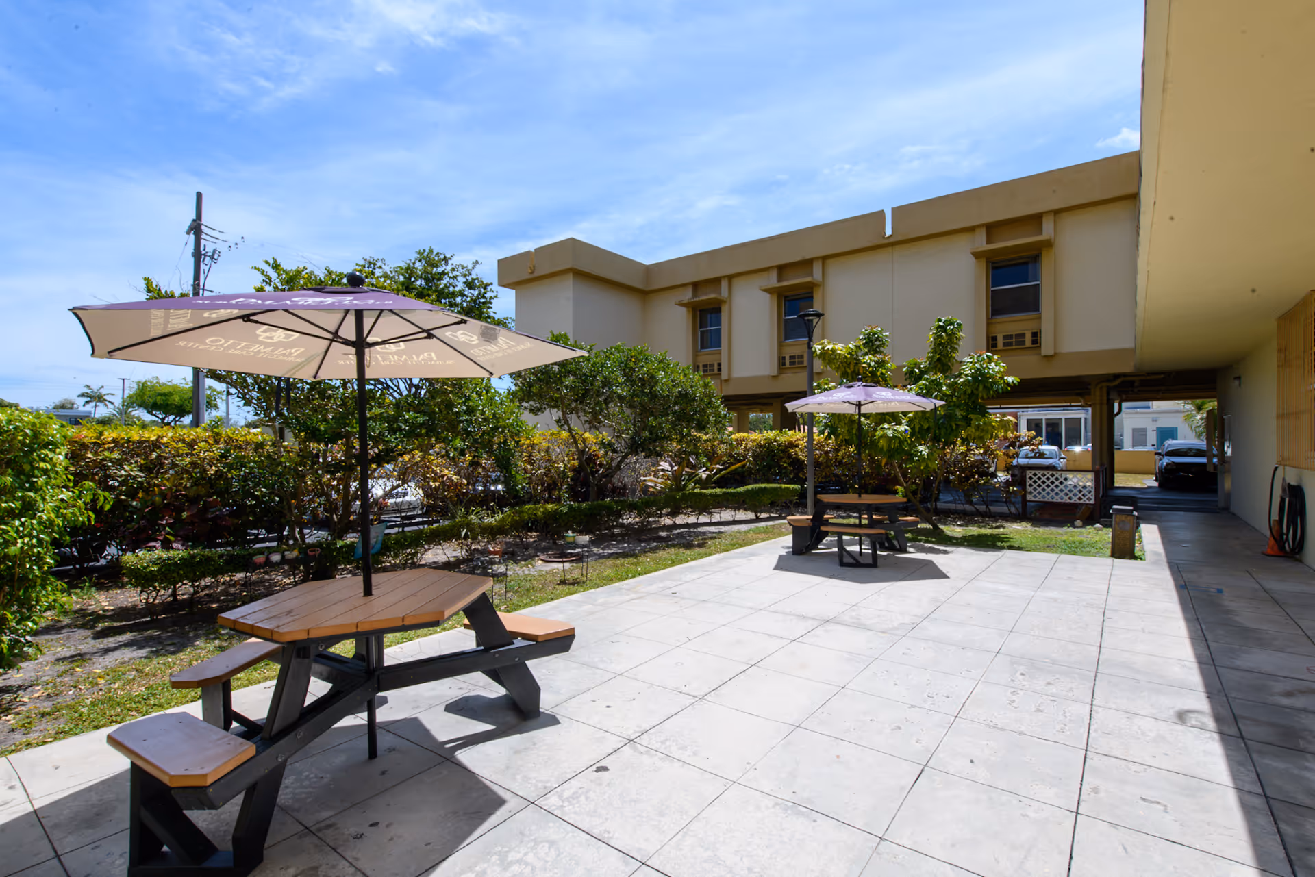 Outdoor patio area at Palmetto Subacute Care Center with two wooden picnic tables, each shaded by a large purple umbrella. The patio is tiled and surrounded by green bushes and trees, with a beige two-story building in the background under a partly cloudy blue sky.
