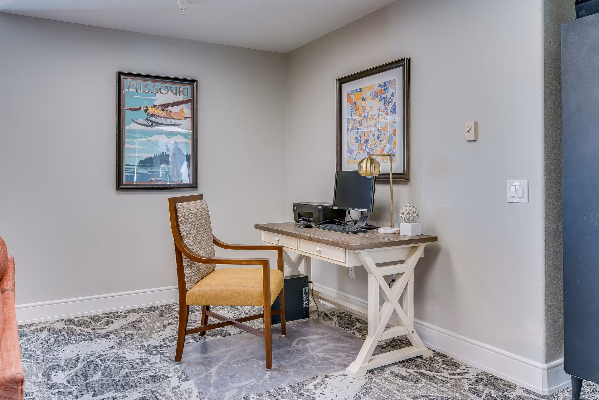 A small office area in a senior living facility with a wooden desk holding a computer, keyboard, printer, and decorative items. A wooden chair with a yellow cushion is positioned in front of the desk. Two framed artworks hang on the light gray walls, and the floor has a patterned carpet with a clear plastic chair mat under the chair.