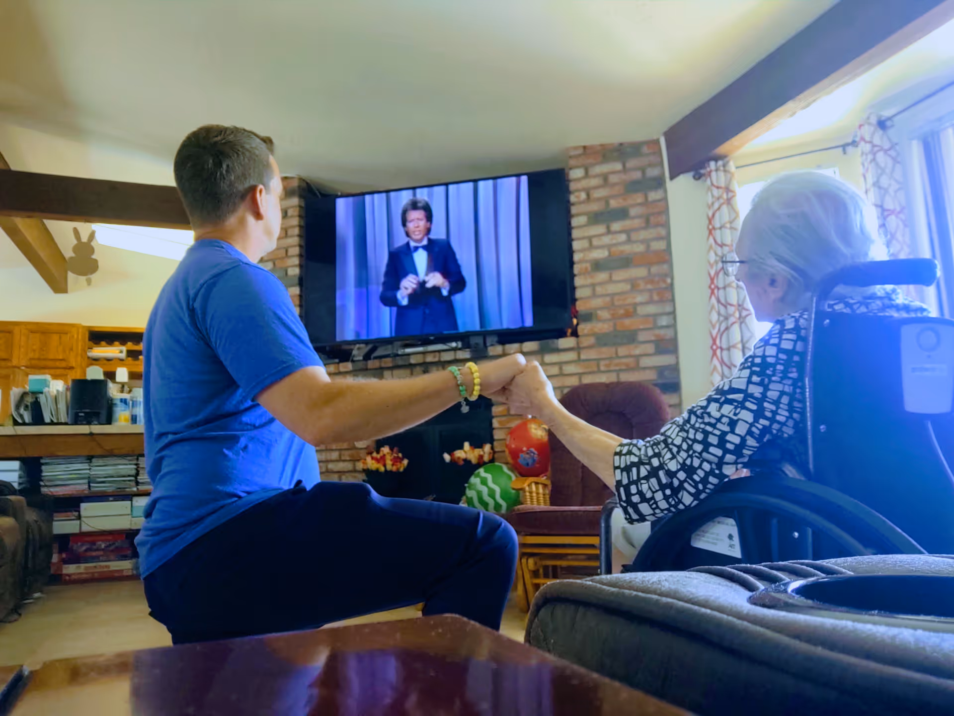 A caregiver holds hands with an elderly woman in a wheelchair while they watch TV in a cozy living room with a brick fireplace.