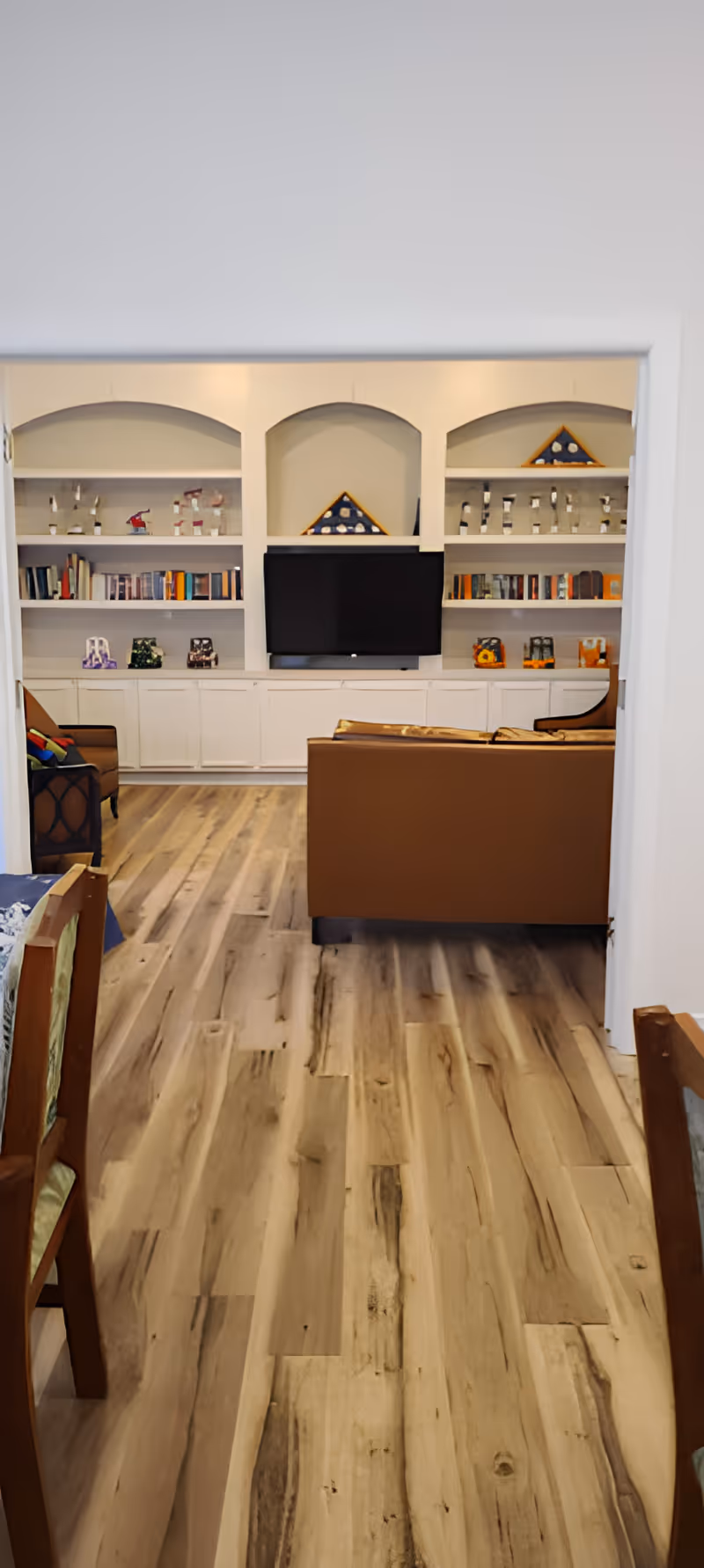 View of a living room area with wooden flooring, a brown sofa, an armchair, and built-in white shelves filled with books and decorative items. A flat-screen TV is mounted in the center of the shelves.