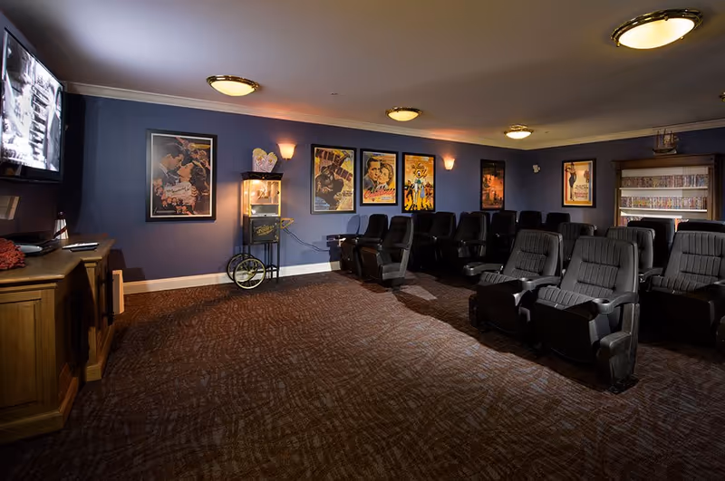 A cozy movie theater room with multiple black reclining chairs arranged in rows facing a wall-mounted TV screen. The walls are painted blue and decorated with framed vintage movie posters. A popcorn machine is placed against the wall, and there is a wooden cabinet below the TV. The room is softly lit with ceiling lights and wall sconces.