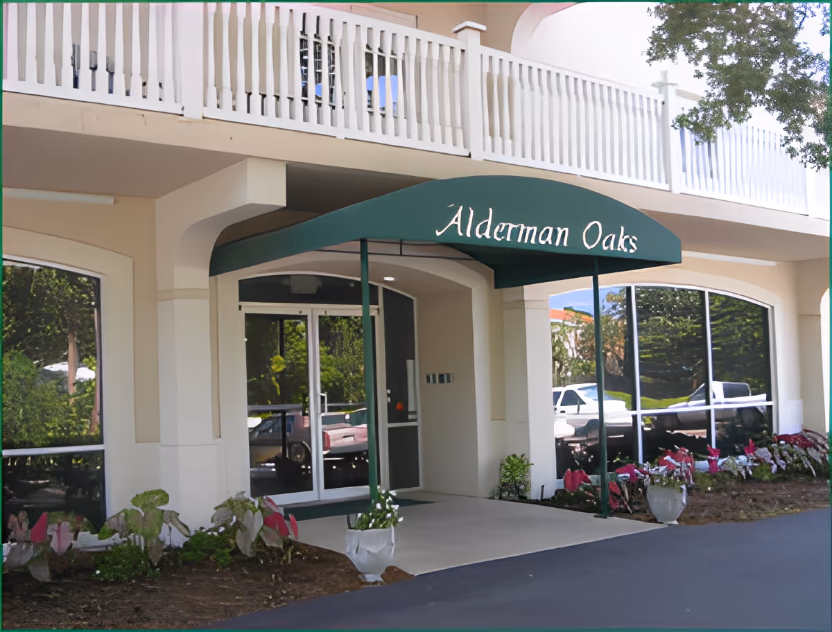 Entrance of Alderman Oaks Retirement Residence featuring a green awning with the facility name, glass double doors, large windows reflecting the outdoor surroundings, and landscaped flower beds on either side of the entrance.