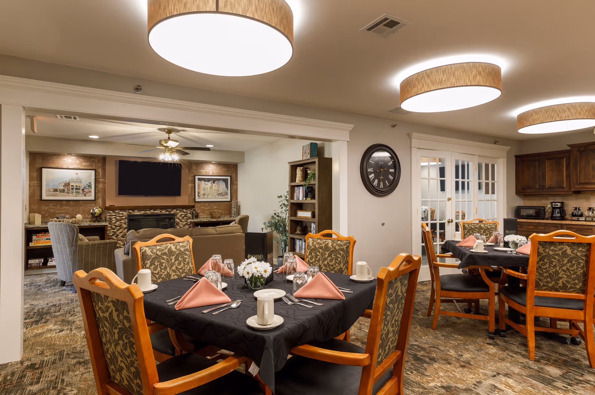 A dining area in a senior living facility with two tables set with black tablecloths, pink folded napkins, white cups, and silverware. The room has wooden chairs with patterned upholstery, modern ceiling lights, and a wall clock. In the background, there is a cozy living room area with a fireplace, a TV mounted above it, framed artwork, and comfortable seating.