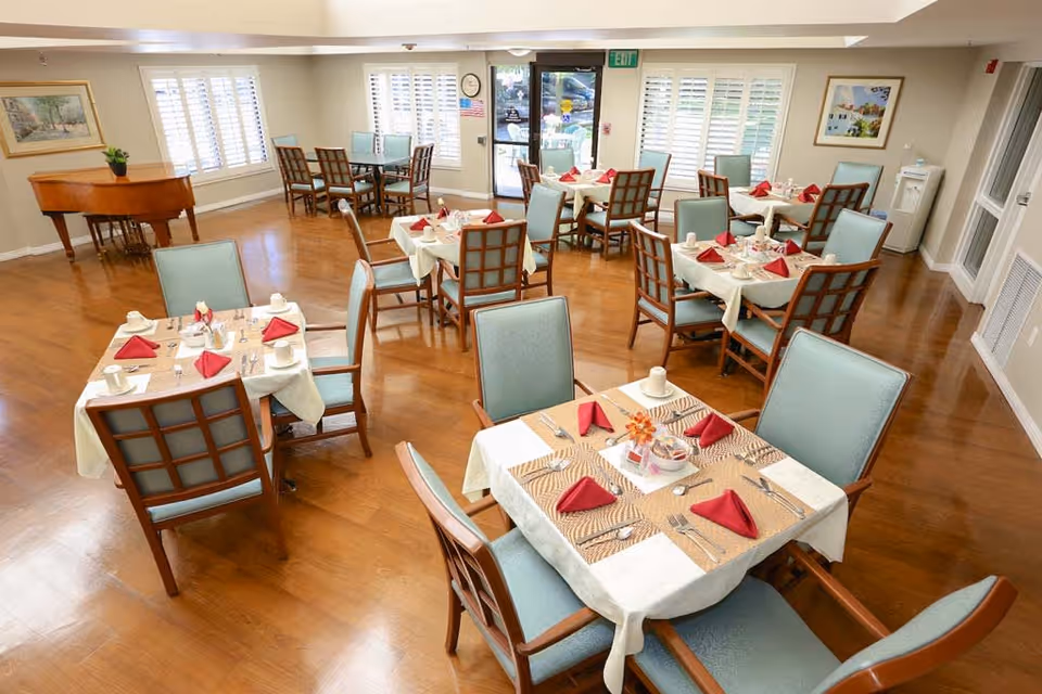 A bright dining room with several square tables covered with white and beige tablecloths, each set with red folded napkins, cups, and silverware. The room has wooden floors, light blue cushioned chairs, large windows with white shutters, a piano in the corner, and framed artwork on the walls.