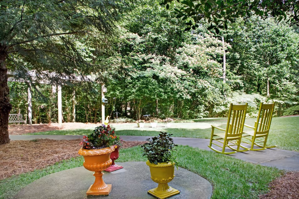 A peaceful outdoor garden area with two yellow rocking chairs on a path, colorful planters on a circular patio, and trees in the background.